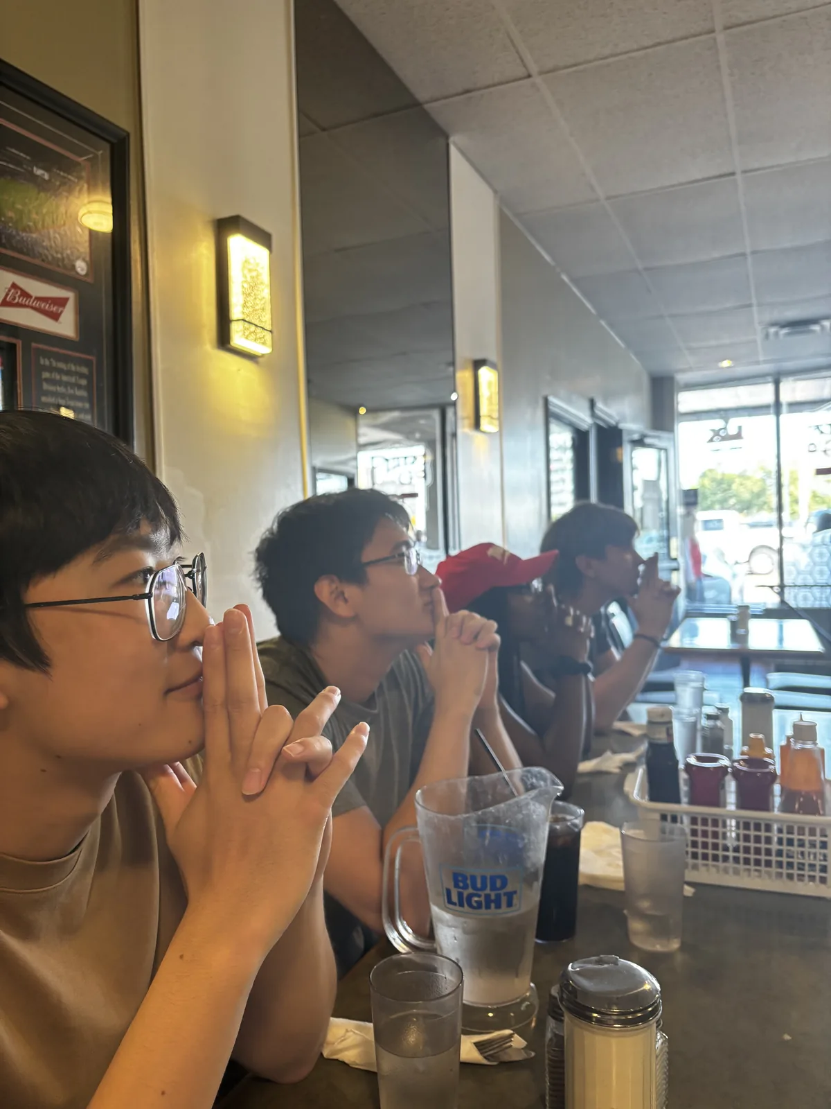 A row of us leaning on a diner counter with hands clasped, watching something off to the side, a pitcher between us