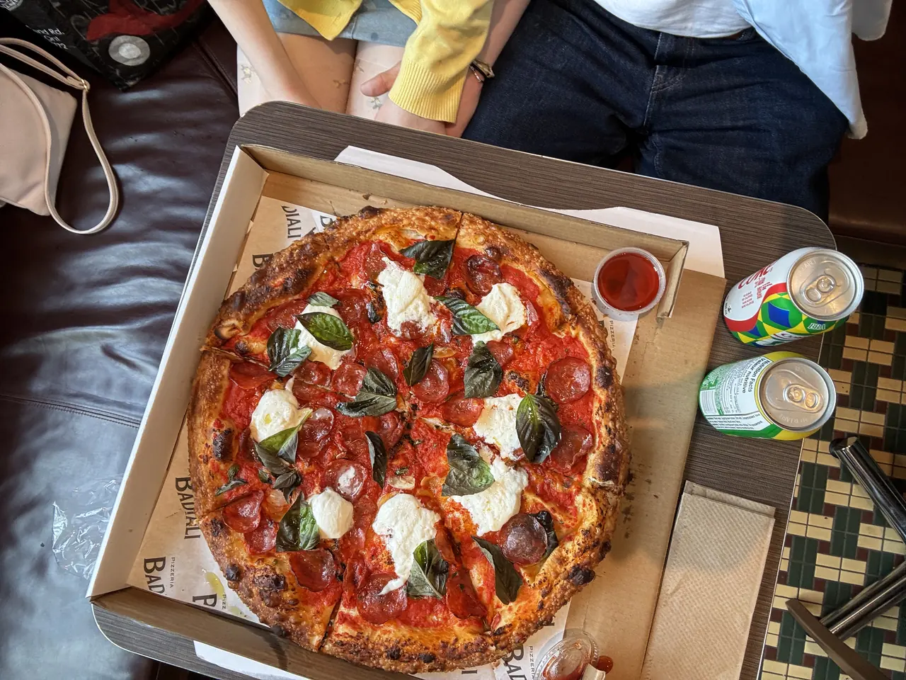 A pepperoni and basil pizza with torn burrata, still in the box, cans of pop beside it