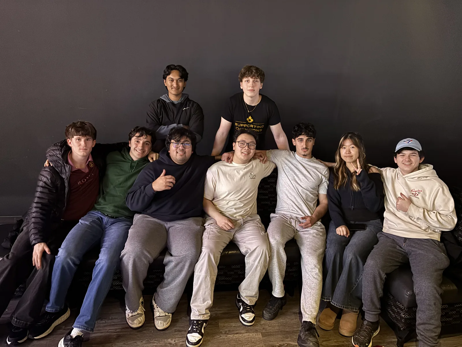 Nine friends packed onto one couch against a dark wall, arms over each other's shoulders